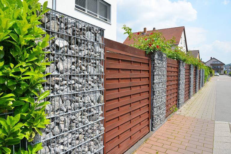 Moderner Sichtschutzzaun aus mit Naturstein gefüllten Gabionen, Holzbretterverschalung und eingegrüntem verzinktem Stahlgitter um ein bebautes Grundstück